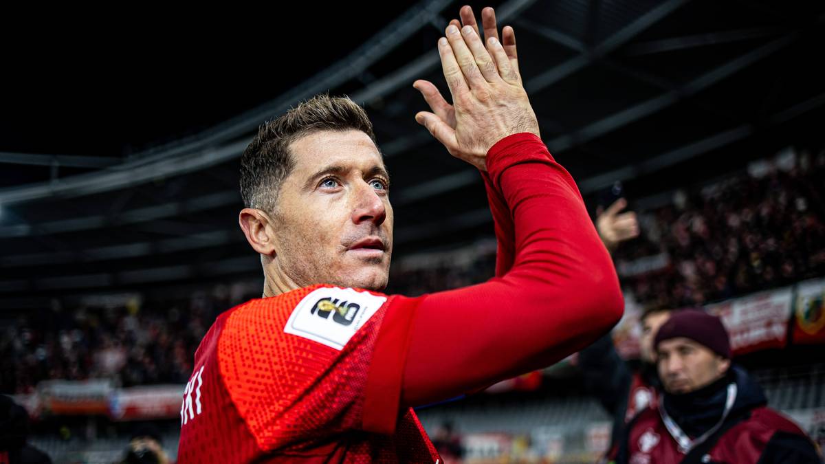 Robert Lewandowski w czerwonej koszulce z numerem i herbem klubu, klaszcze z uniesionymi dłońmi, na tle trybun stadionu.