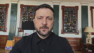 Prezydent Ukrainy Wołodymyr Zełenski w czarnym ubraniu przemawia do kamery, zza jego plec&oacute;w widoczne są ozdobne ściany z ramkami.