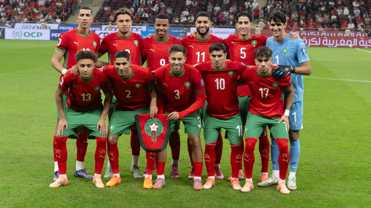 Drużyna piłkarska Maroka w czerwonych strojach pozuje do zdjęcia na boisku, z herbem Maroka na tarczy.
