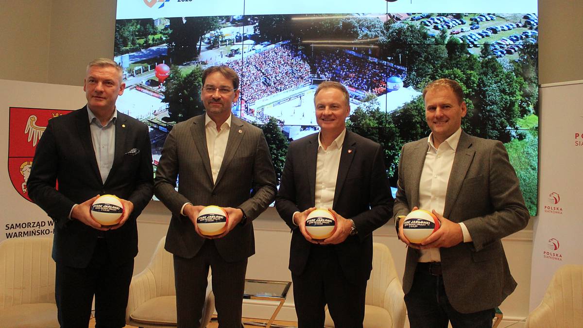 Czterech mężczyzn w garniturach trzymających piłki do siatkówki przed ekranem z widokiem stadionu.