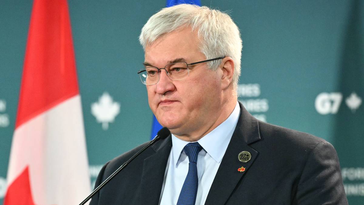 Mężczyzna w okularach i garniturze stoi za mównicą z mikrofonem, w tle widoczne flagi Kanady i G7.