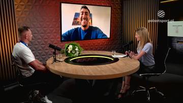 Uśmiechnięty mężczyzna na ekranie podczas wywiadu w studio telewizyjnym z dwiema osobami siedzącymi przy okrągłym stole.