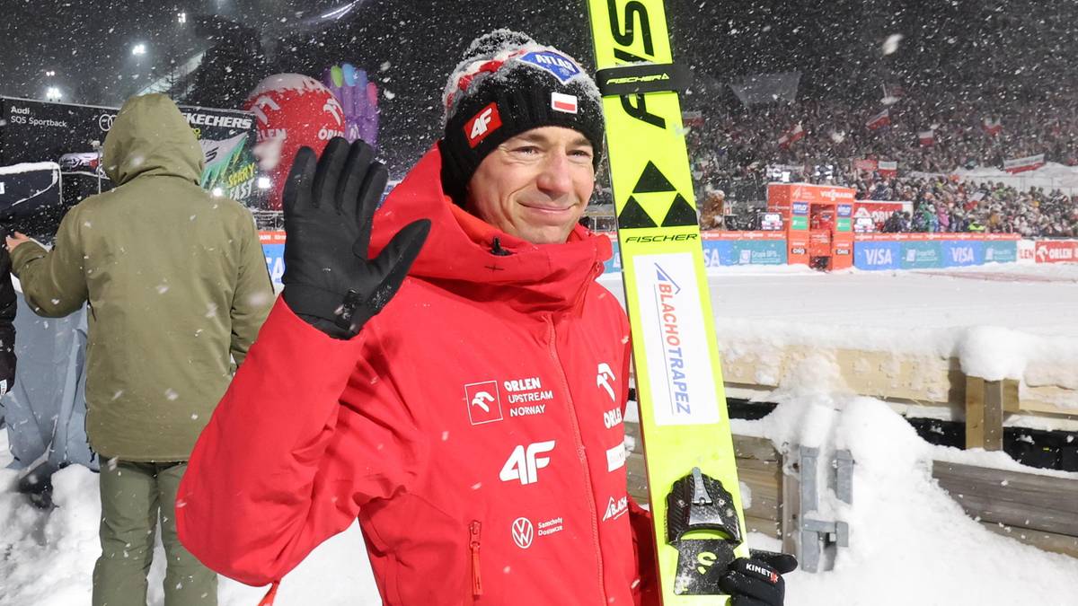 Skoczek narciarski w czerwonej kurtce z polskim orłem na rękawie, trzyma nartę z żółtym oznaczeniem i macha do publiczności.