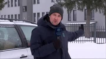 Dziennikarz w zimowym ubraniu trzyma mikrofon i wskazuje ręką w stronę budynku podczas relacji na śnieżnym tle.