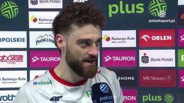 Portret mężczyzny z brodą i kręconymi włosami, ubranego w białą koszulkę sportową z czerwonym wykończeniem, podczas wywiadu na tle ścianki sponsorskiej z logo "Plus" i "Orlen".