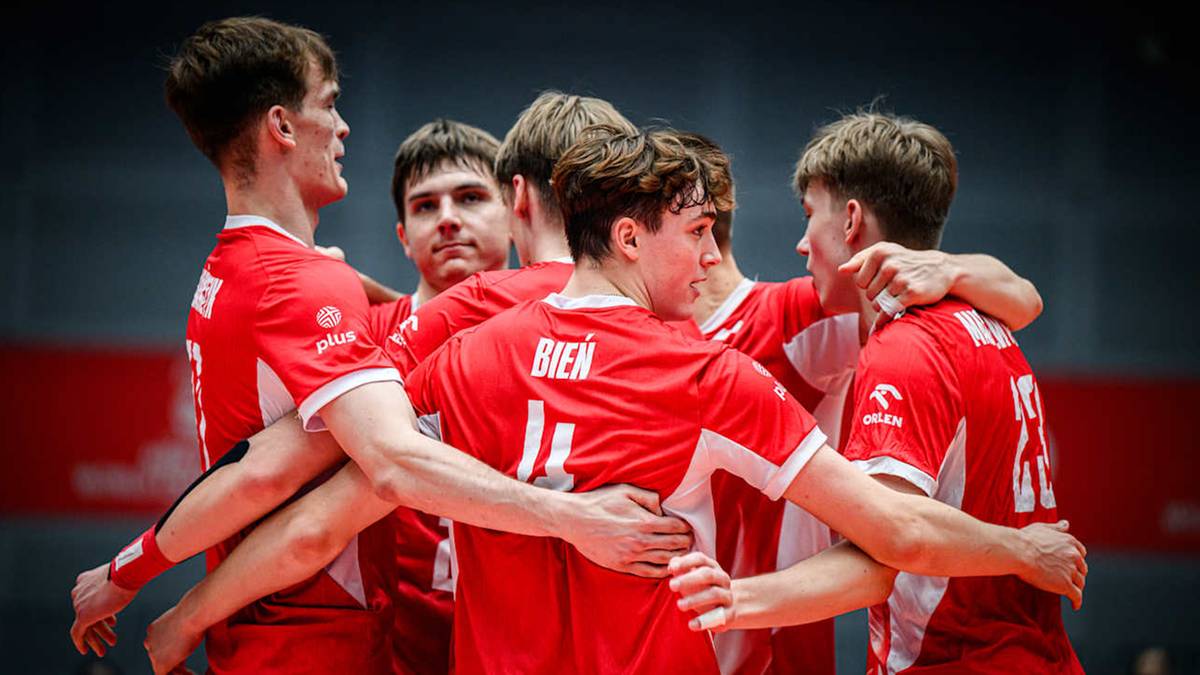 Drużyna siatkarzy w czerwonych strojach okrąża się, okładając się po plecach, patrząc w kierunku kamery.