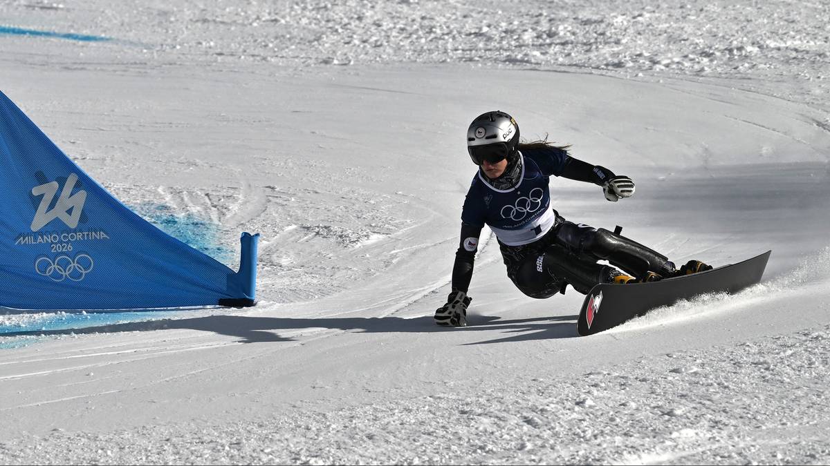 Snowboarderka w kasku i ciemnym stroju sportowym zjeżdża po ośnieżonym stoku, wykonując skręt. Na niebieskim banerze w tle widoczne są olimpijskie k&oacute;łka.