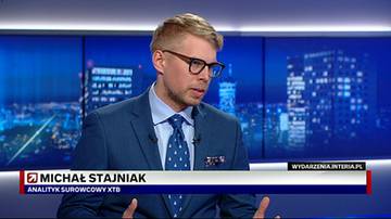 Mężczyzna w granatowym garniturze i okularach patrzy w prawo. Za nim znajduje się ekran z widokiem nocnego miasta i napisem "Michał Stajniak Analityk surowcowy XTB".