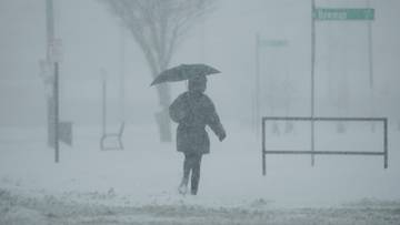 Osoba z parasolką idąca przez zaśnieżoną ulicę podczas silnych opadów śniegu, widoczność jest ograniczona.