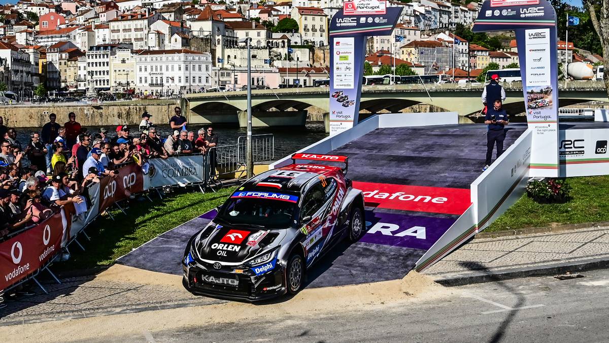 Samoch&oacute;d rajdowy z numerem startowym 2, z logiem LOTOS Rally na masce, wjeżdża na podium, za nim rozmyte tłumy widz&oacute;w i zabudowania miejskie z wzg&oacute;rzem w tle.