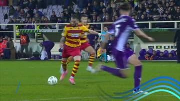 Piłkarz w żółto-czerwonym stroju próbuje odebrać piłkę zawodnikowi w fioletowym stroju, który właśnie kopie piłkę.