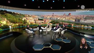 Sześciu mężczyzn siedzi w okręgu na podwyższeniu w studiu telewizyjnym. W tle wirtualne panorama miasta z widokiem na rzekę i wzgórza. Po prawej stronie ekranu widoczna jest młoda kobieta z podniesionymi rękami i otwartymi ustami, prawdopodobnie komentatorka.