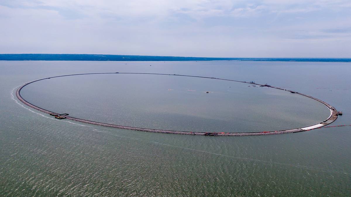 Widok z lotu ptaka na dużą, owalną sztuczną wyspę zbudowaną na Zalewie Wiślanym, otoczoną stalowymi ściankami Larsena.