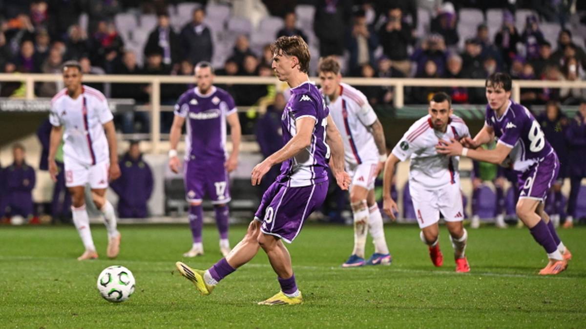 Piłkarz w purpurowym stroju wykonuje rzut wolny, podczas gdy inni zawodnicy w białych i purpurowych strojach obserwują akcję.