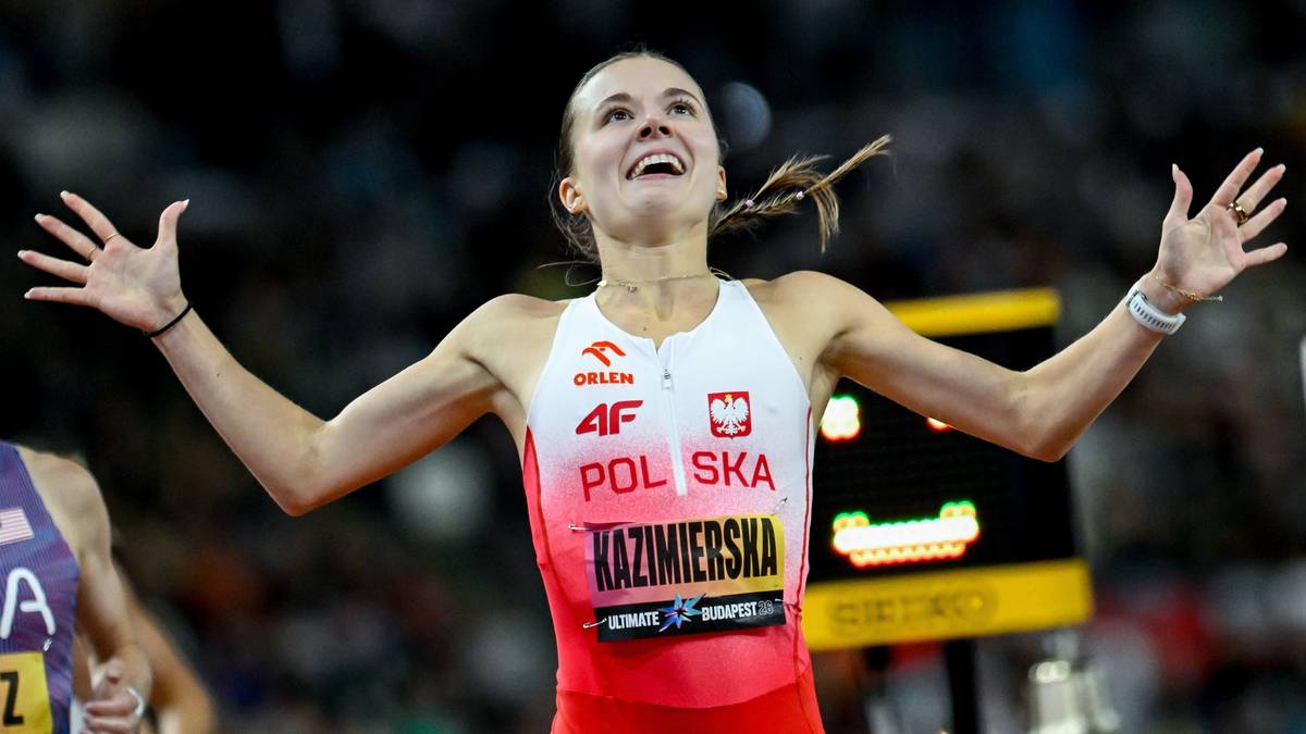 Szczęśliwa lekkoatletka w białej i czerwonej koszulce z napisem "POLSKA" i "KAZIMIERSKA" szeroko rozkłada ramiona w geście triumfu po przekroczeniu linii mety na stadionie.