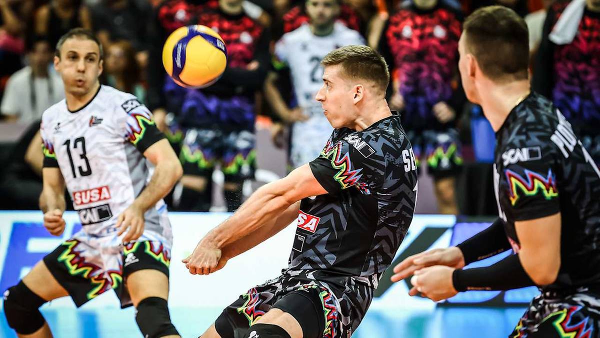 Siatkarz w czarnej koszulce z kolorowymi wzorami odbija piłkę do siatkówki, z zawodnikami w białych i czarnych koszulkach w tle.