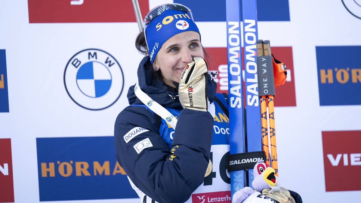 Kobieta w stroju biathlonowym, w niebieskiej czapce z pomponem i w rękawicach, trzyma narty. Na jej twarzy maluje się radość.