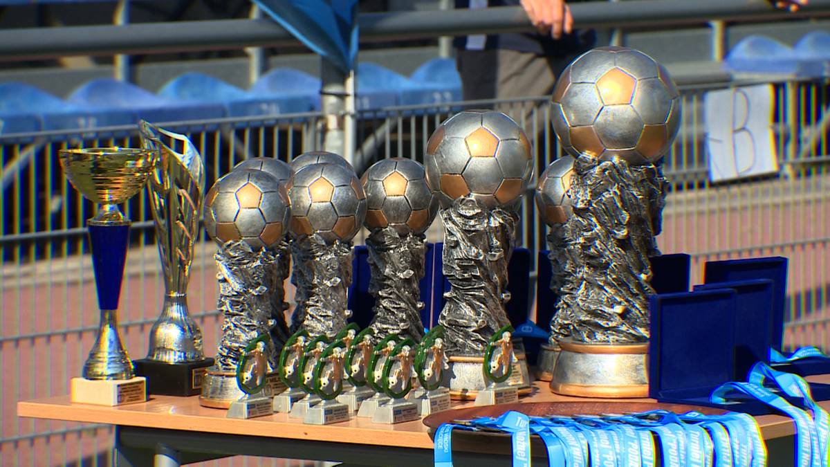 Kilka piłkarskich trofeów, w tym duża statuetka w kształcie piłki na postumencie, umieszczonych na stole. W tle widać niebieskie krzesełka trybun.