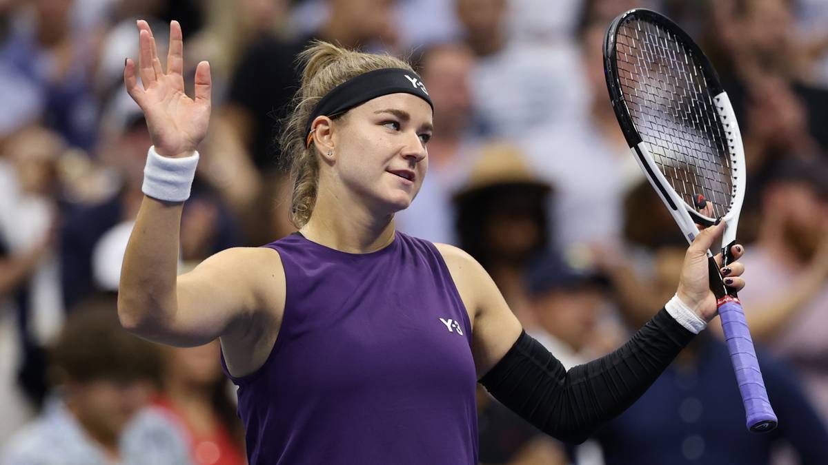 Tenisistka w fioletowej koszulce sportowej, z czarną opaską na głowie i białym frotte na lewym nadgarstku, podnosi prawą rękę na wysokość głowy, machając dłonią, a w lewej trzyma rakietę tenisową.