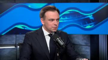 Mężczyzna w ciemnym garniturze i białej koszuli z krawatem przemawia do mikrofonu. Za nim znajduje się niebieski ekran z abstrakcyjnymi, falistymi wzorami.