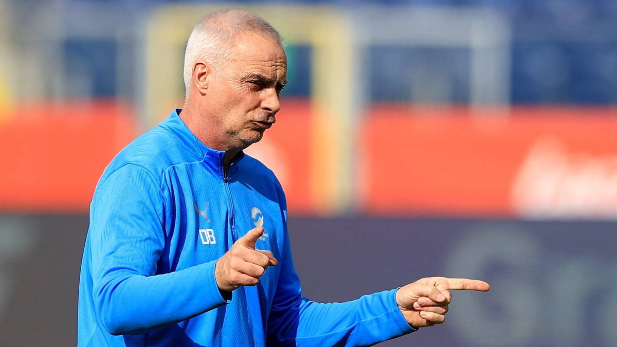 Mężczyzna w niebieskiej kurtce sportowej, prawdopodobnie trener piłkarski, wskazuje palcami w bok.