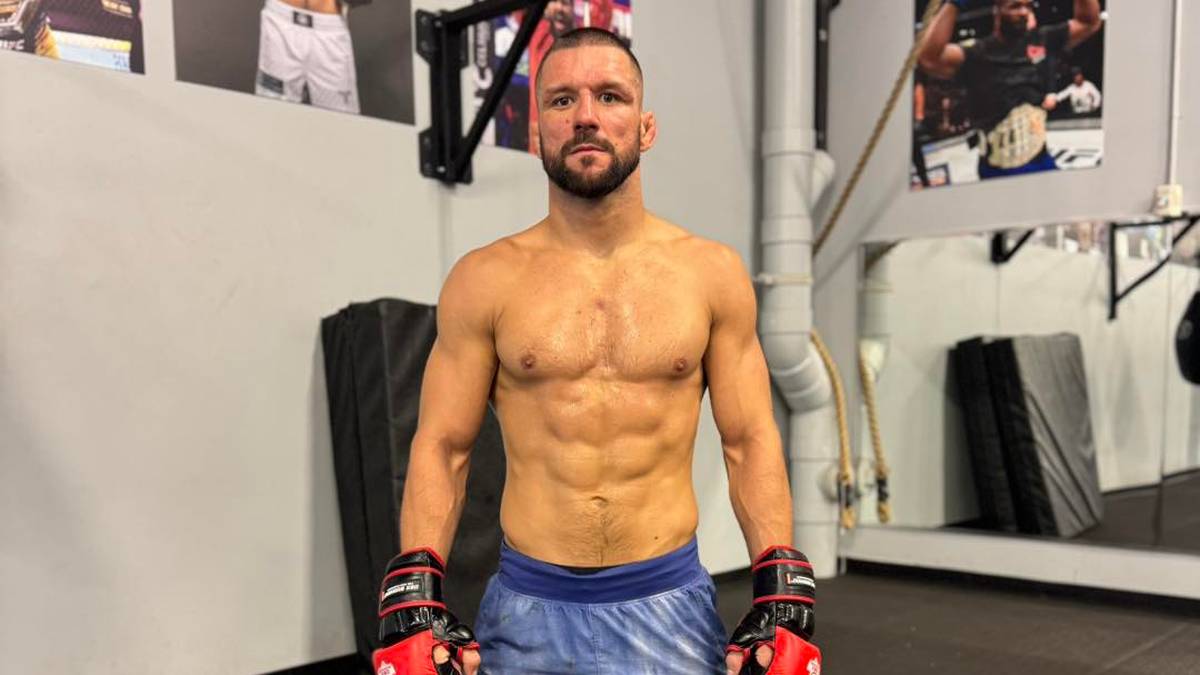 Mateusz Gamrot w stroju do sport&oacute;w walki, w rękawicach i ochraniaczach na piszczele, pozuje na tle sali treningowej.