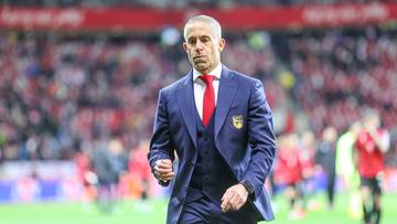 Sylvinho, trener reprezentacji Albanii, w garniturze na boisku piłkarskim.