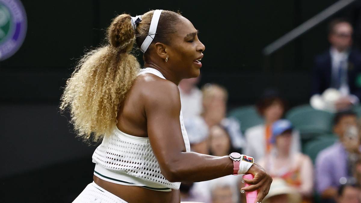Serena Williams klęczy na kortach Wimbledonu z rakietą obok siebie, podczas gdy tłum kibiców siedzi na trybunach w tle.