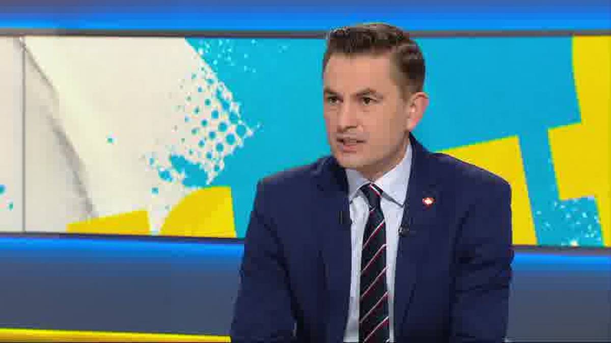 Mężczyzna w granatowym garniturze, białej koszuli i krawacie siedzi na tle studia telewizyjnego z kolorowym, geometrycznym motywem.