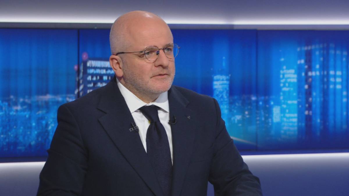 Dojrzały mężczyzna w okularach, w garniturze i krawacie, siedzi przed ekranem z widokiem na nocne miasto.