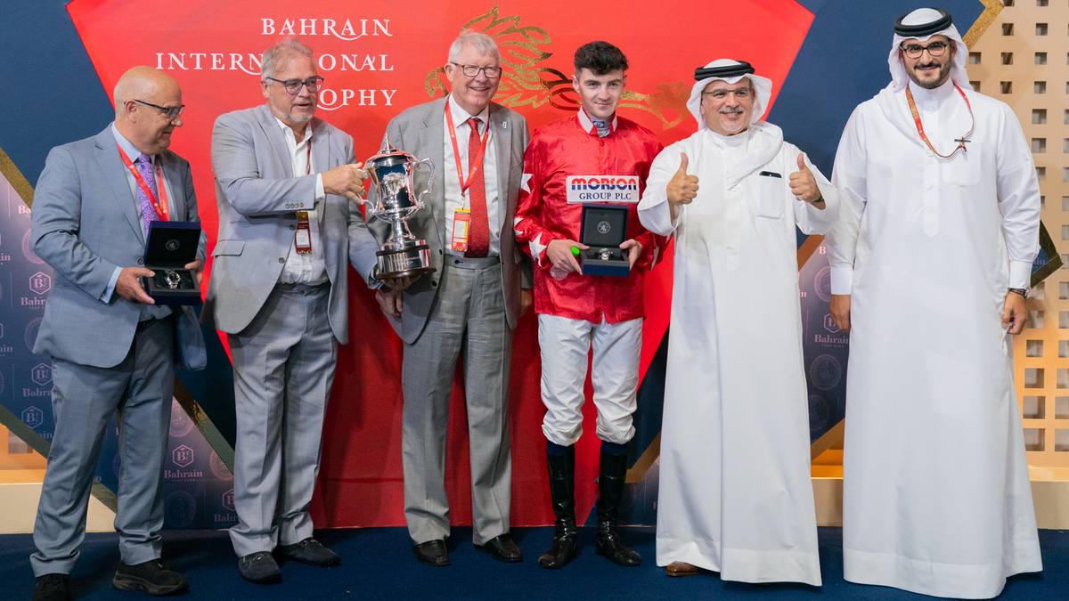 Mężczyźni w eleganckich strojach i dżokej pozują z pucharami na tle sceny Bahrain International Trophy.