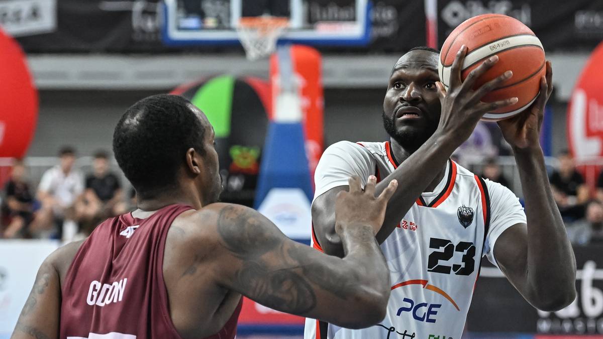 Dwóch koszykarzy podczas meczu. Jeden zawodnik w białej koszulce z numerem 23 na plecach trzyma piłkę do koszykówki, przygotowując się do rzutu. Drugi zawodnik w bordowej koszulce stara się go zablokować.