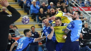 Zawodnicy piłki ręcznej walczą o piłkę podczas meczu, jeden gracz w niebieskim stroju z numerem 34 pr&oacute;buje przechwytu.