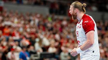 Piłkarz ręczny w białej koszulce z czerwonymi elementami krzyczy podczas meczu, z publicznością na trybunach w tle.