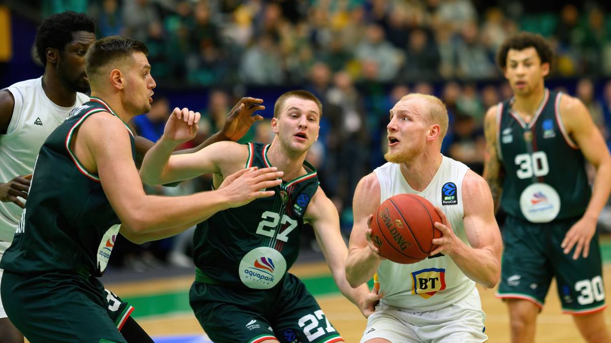 Czterech zawodników drużyny koszykarskiej walczy o piłkę. Dwóch w białych strojach i dwóch w zielonych. Na dalszym planie widać zamazane sylwetki kibiców na trybunach.