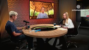 Dw&oacute;ch młodych ludzi siedzi przy okrągłym stole w studiu telewizyjnym, rozmawiając z mężczyzną widocznym na ekranie.