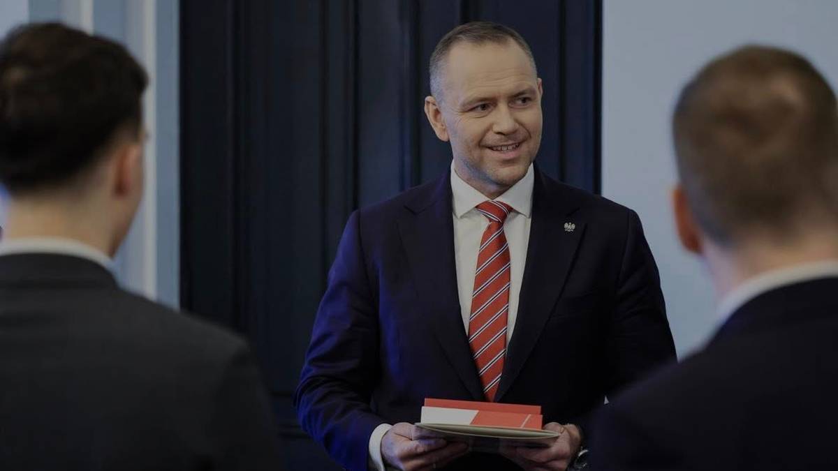 Karol Nawrocki w garniturze i krawacie trzyma w rękach dokumenty, zwraca się do rozm&oacute;wc&oacute;w.