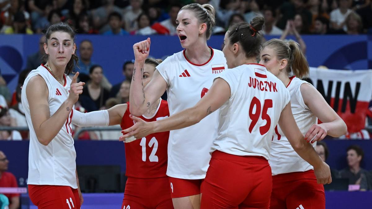 Pięć zawodniczek w białych koszulkach z czerwonymi elementami i numerami na plecach, otoczonych przez innych ludzi na trybunach. Jedna z zawodniczek jest na pierwszym planie, z podniesioną ręką w geście radości. Widoczny jest napis "JUSZCZYK 95" na plecach jednej z zawodniczek.