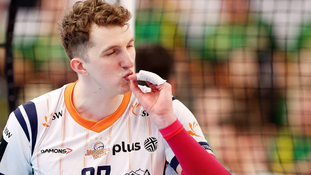 Mężczyzna w koszulce sportowej z numerem 27, wkładający coś do ust, z siatką do siatk&oacute;wki w tle.