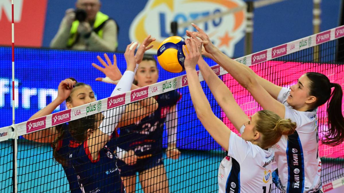 Moment zagrania w siatkówce, cztery zawodniczki próbują zablokować piłkę nad siatką podczas meczu. Jedna z nich dotyka piłki ręką.