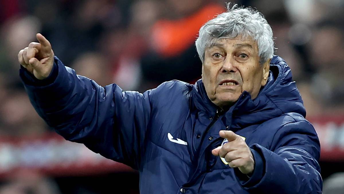 80-letni trener piłkarski w niebieskiej kurtce z białym logo wskazuje palcem w bok, z wyraźnym wyrazem twarzy. W tle widać trybuny stadionu.