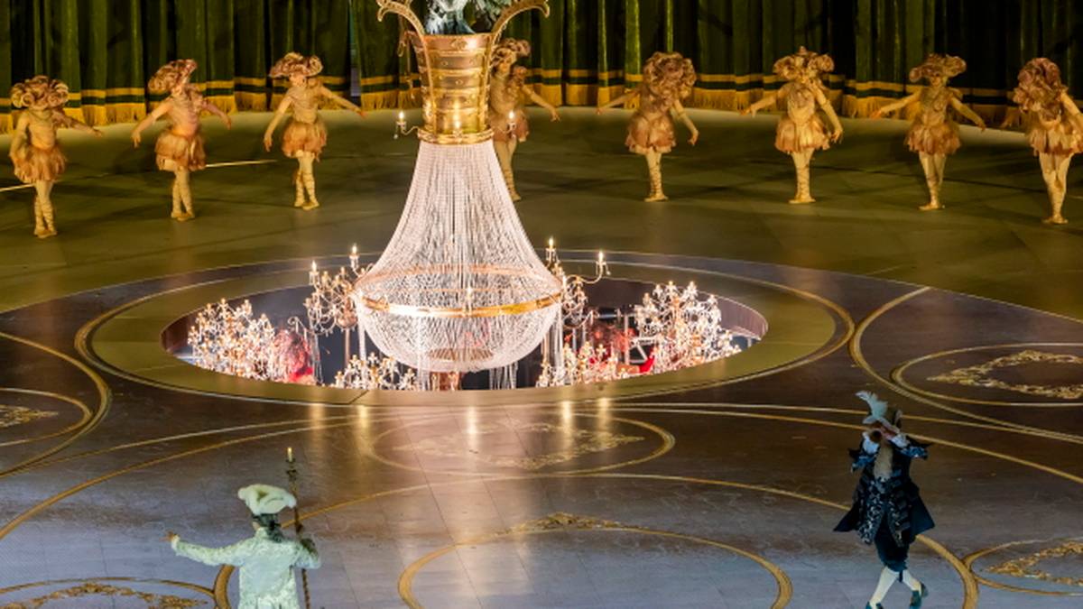 Scena z występem artystycznym z centralnie umieszczonym żyrandolem i grupą tancerzy w strojach scenicznych.