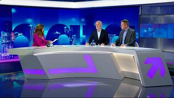 Trzy osoby siedzące przy nowoczesnym, zakrzywionym stole w studiu telewizyjnym. Po lewej kobieta, po prawej dwaj mężczyźni w garniturach. Wszyscy są skierowani w tym samym kierunku, sugerując dyskusję lub wywiad.