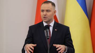 Mężczyzna w garniturze przemawia przy mikrofonie, za nim widoczne są flagi Polski i Ukrainy.