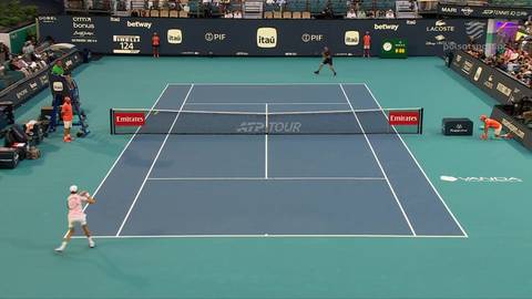 Tenisista w białej koszulce i r&oacute;żowych spodenkach odbija piłkę na niebieskim korcie tenisowym. W tle widać drugiego tenisistę, siatkę i trybuny z widzami.