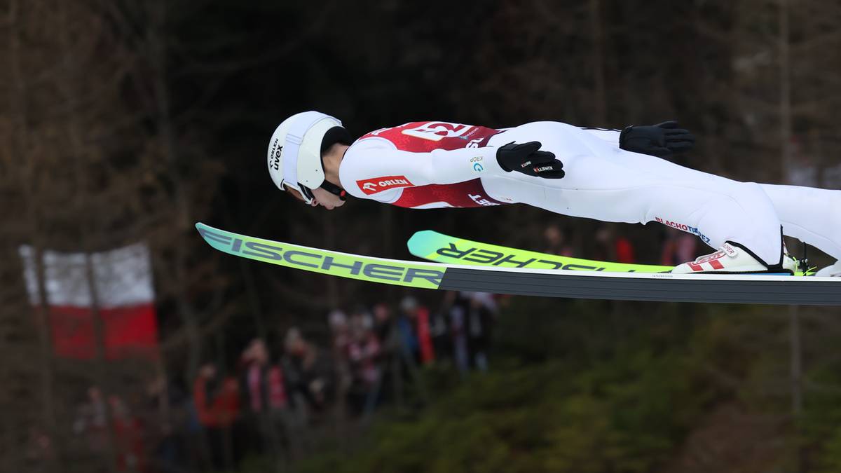 Skoczek narciarski w locie, ubrany w biało-czerwony kombinezon i biały kask, z nartami Fischer.