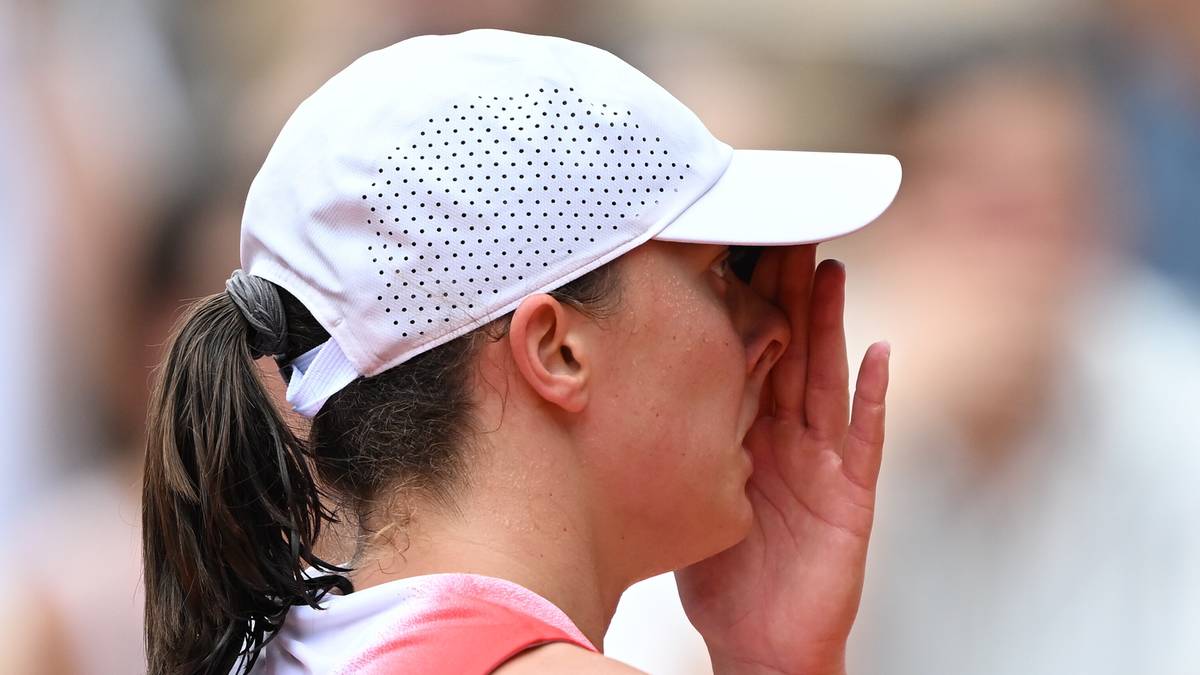 Tenisistka w białej czapce z daszkiem dotyka dłonią twarzy, patrząc w bok. Jej ciemne włosy związane są w kucyk. Tło jest rozmyte.