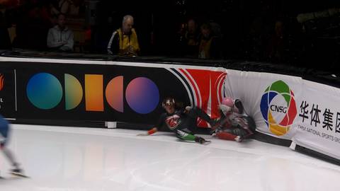 Dwóch zawodników z polskiej sztafety upadło na lodowisku podczas zawodów panczenowych. Jeden z nich leży na plecach, drugi na boku, obaj w kombinezonach sportowych. W tle widoczni są inni panczeniści oraz fragmenty band reklamowych z logotypami.