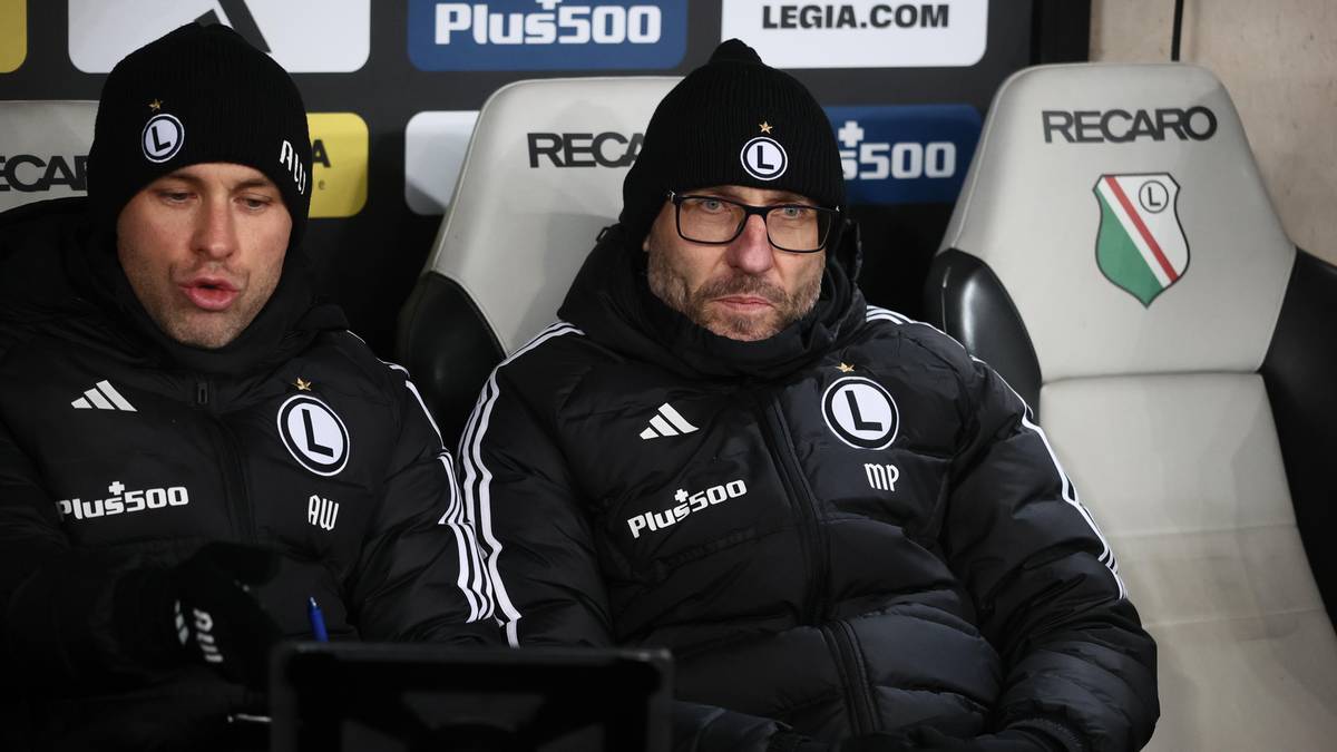 Dwóch mężczyzn w czapkach i kurtkach z logo Legii Warszawa siedzi na ławce rezerwowych podczas meczu.