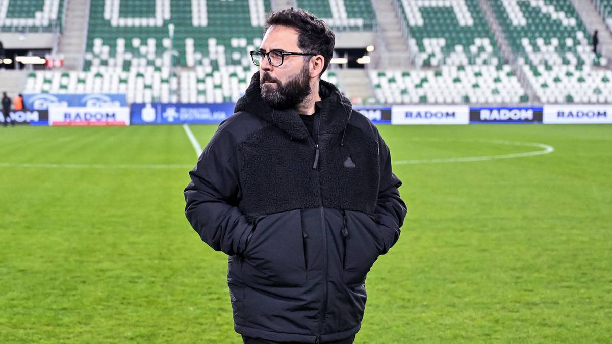 Mężczyzna w kurtce stoi na boisku piłkarskim przed trybunami stadionu z napisem Radomiak.